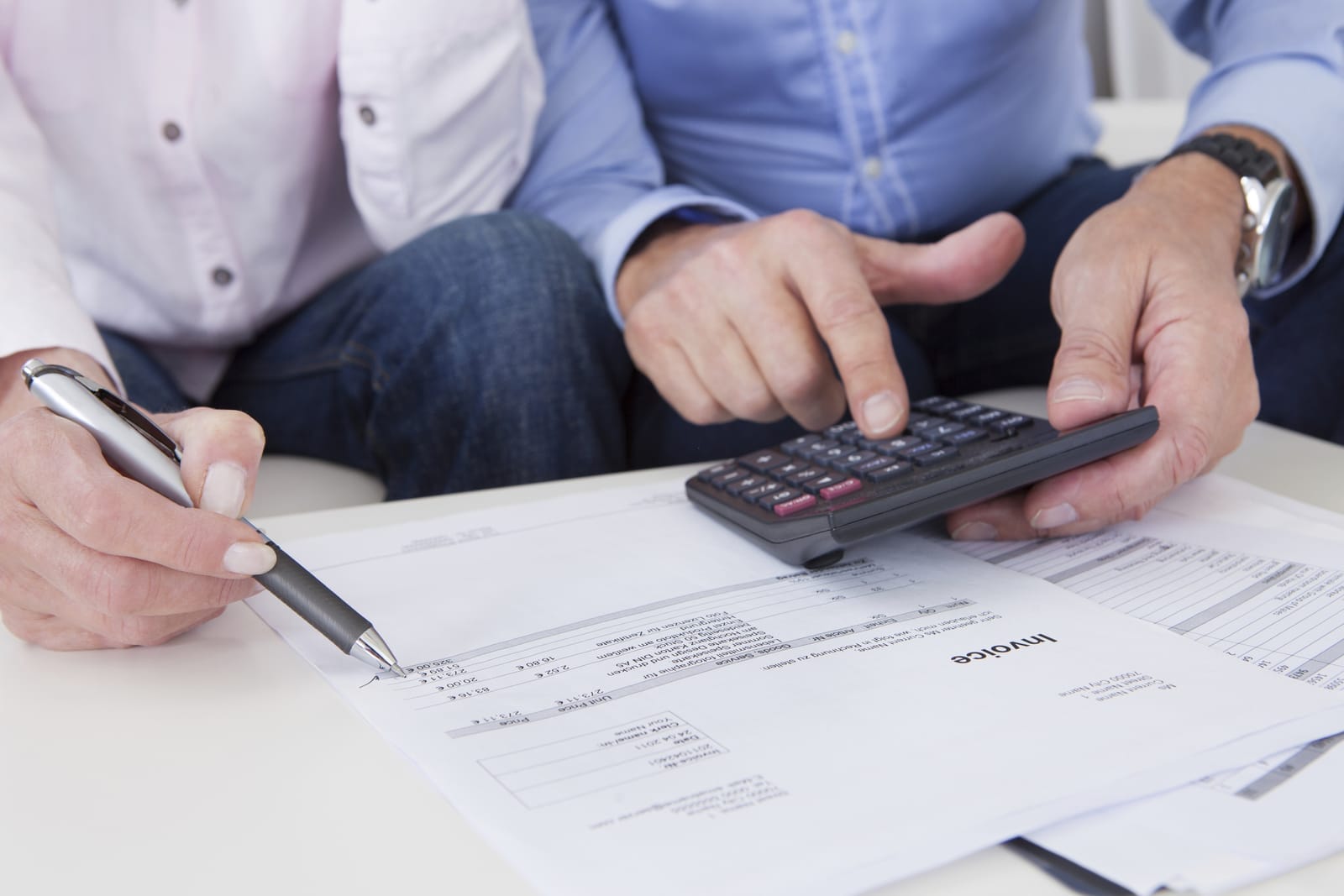 Two people reviewing invoices; one holds a pen, the other uses a calculator. Papers cover a table, with a focused and collaborative mood.