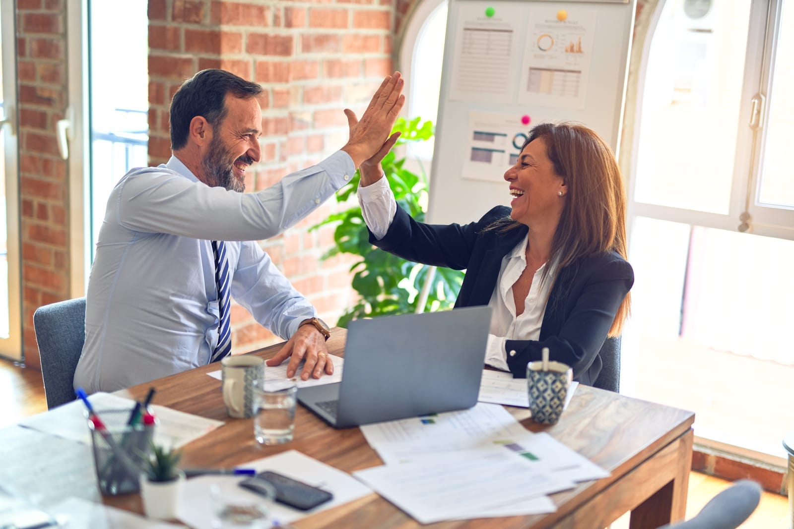 Two colleagues high-five at a wooden table in an office. Papers, laptop, and mugs are visible. Bright daylight through large windows. Happy mood.